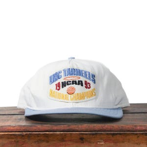 May include: White baseball cap with a light blue brim. The cap features an embroidered patch with the text "UNC Tar Heels 19 NCAA 93 National Champions" in blue, red, and gold.