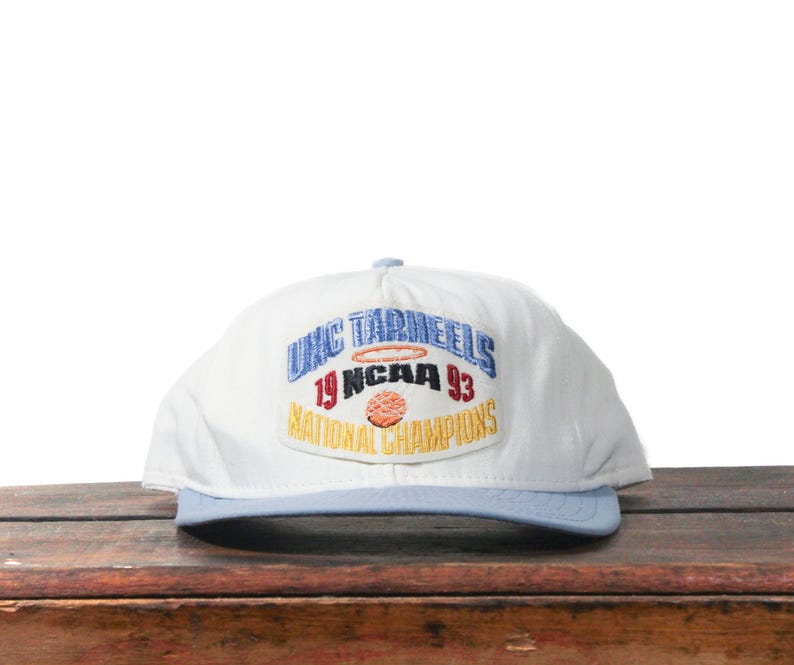 May include: White baseball cap with a light blue brim. The cap features an embroidered patch with the text "UNC Tar Heels 19 NCAA 93 National Champions" in blue, red, and gold.