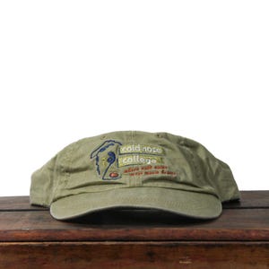 May include: Olive green baseball cap with the embroidered text "cold nose college" and a cartoon graphic. The cap is made of a soft, worn material and sits on a wooden surface.