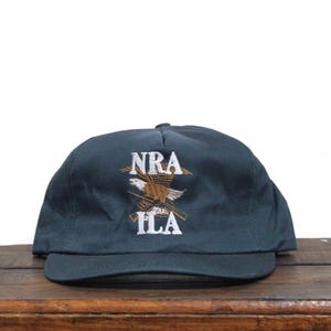 以下が含まれることがあります： 濃い青色のベースボールキャップに、刺繍されたロゴが施されています。ロゴは「NRA」の文字と、その下に交差したライフルを持ったワシが描かれています。ロゴは白と茶色です。