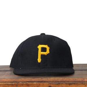 Könnte beinhalten: Schwarze Baseballkappe mit einem gelb gestickten "P" auf der Vorderseite. Die Kappe hat einen flachen Schirm und eine runde Krone. Der Hut liegt auf einer Holzoberfläche.