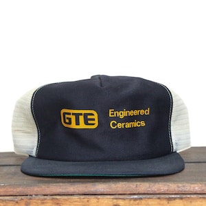 May include: A navy blue trucker hat with a white mesh back and a black brim. The front panel displays the yellow logo "GTE" and the text "Engineered Ceramics" in yellow.