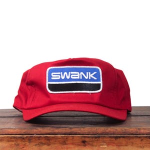 May include: A red baseball cap with a rectangular patch displaying the word "SWANK" in white text on a blue background, outlined in black and white. The cap is on a wooden surface.