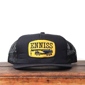 May include: Black trucker hat with a yellow patch featuring the word "ENNISS" and a crane graphic. The hat has a mesh back and a curved brim. The hat is sitting on a wooden surface.