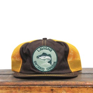 Peut inclure: Casquette de camionneur marron et jaune avec un patch vert et blanc qui dit "Lavigne Ontario Canada" et représente un poisson.