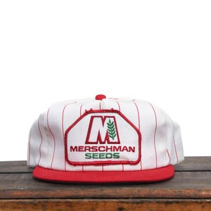 Peut inclure: Casquette de baseball blanche à rayures rouges et un patch brodé rouge, blanc et vert qui dit "Merschman Seeds" avec un grand "M" et une tige de blé verte.