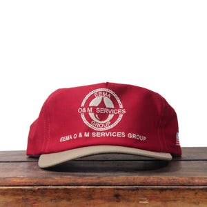 May include: A red baseball cap with a tan brim. The cap features white embroidered text and a circular logo that reads "EEMA O&M SERVICES GROUP". The cap is sitting on a wooden surface.
