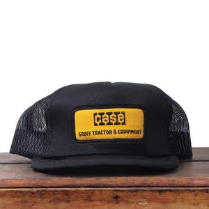 Op de afbeelding: Zwarte truckerpet met een gele rechthoekige patch. De patch toont het woord "CASE" in een vetgedrukte lettertype boven "GROFF TRACTOR & EQUIPMENT". De pet heeft een mesh achterkant.