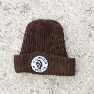 以下が含まれることがあります： 黒い文字で「R.C. Musson Rubber Co.」と書かれた白い円形のワッペンが付いた、茶色のニットビーニー。