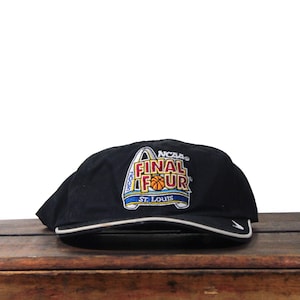 May include: Black baseball cap with the text "NCAA FINAL FOUR ST. LOUIS" in a colorful design. The cap has a white trim and a curved bill. The cap is sitting on a wooden surface.