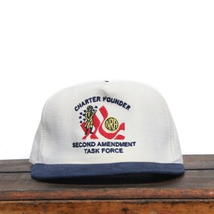 Op de afbeelding: Witte baseballpet met een blauwe rand. De voorkant van de pet heeft een geborduurd logo met de tekst "Charter Founder" en "Second Amendment Task Force". Het logo bevat ook een afbeelding van een persoon die een geweer vasthoudt en het NRA-logo.