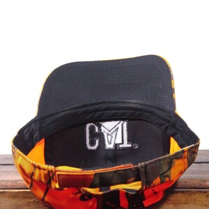 May include: A camouflage baseball cap with a black interior and a brown leather strap. The cap features the embroidered letters "CYL" in white on the inside. The exterior has a mix of orange, yellow, and green colors.