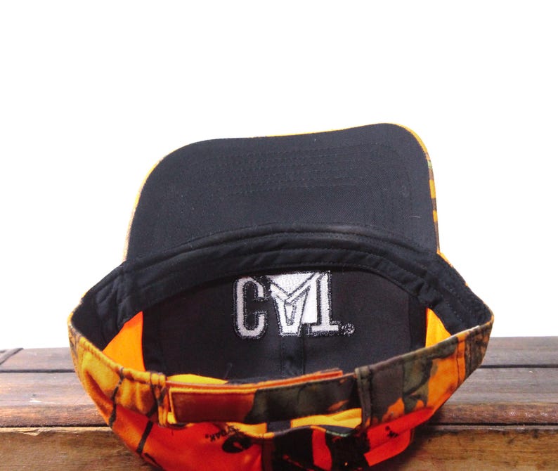 May include: A camouflage baseball cap with a black interior and a brown leather strap. The cap features the embroidered letters "CYL" in white on the inside. The exterior has a mix of orange, yellow, and green colors.