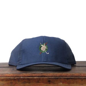 May include: A navy blue baseball cap with a curved brim. The cap features an embroidered design of a golf club and a leaf inside a green circle. The cap is sitting on a wooden surface.