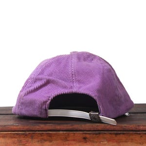 May include: A purple corduroy baseball cap with a curved brim. The cap has a silver-colored adjustable strap and buckle. The hat is sitting on a wooden surface.