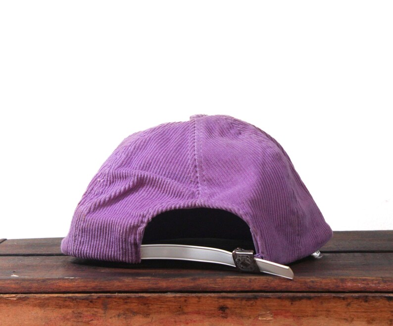 May include: A purple corduroy baseball cap with a curved brim. The cap has a silver-colored adjustable strap and buckle. The hat is sitting on a wooden surface.