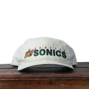 May include: An off-white baseball cap featuring the embroidered text SEATTLE SONICS in dark green and red. To the left of the text is a small, colorful embroidered logo depicting a basketball with a stylized graphic design.
