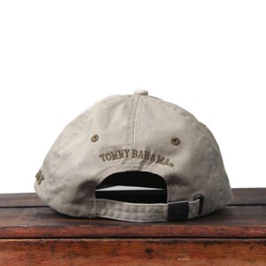 Może przedstawiać: Beżowa czapka z daszkiem z haftowanym napisem "TOMMY BAHAMA" z tyłu. Czapka ma zakrzywiony daszek i regulowany pasek. Czapka leży na drewnianej powierzchni.