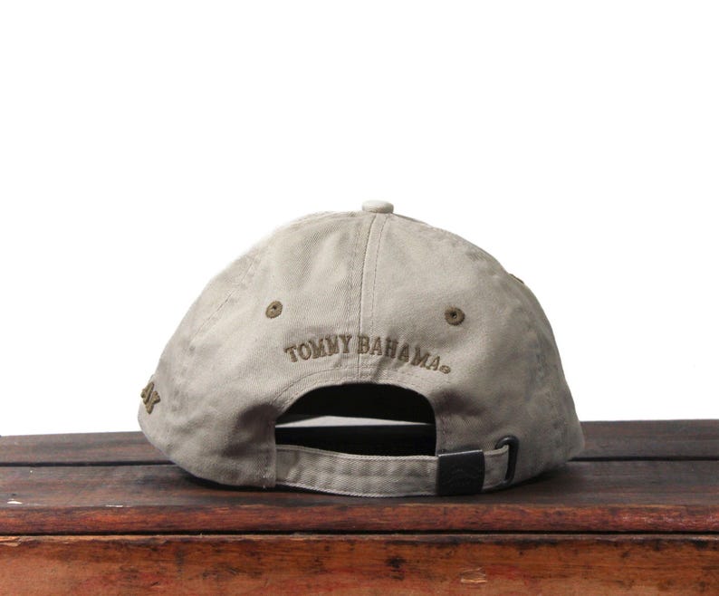 Może przedstawiać: Beżowa czapka z daszkiem z haftowanym napisem "TOMMY BAHAMA" z tyłu. Czapka ma zakrzywiony daszek i regulowany pasek. Czapka leży na drewnianej powierzchni.