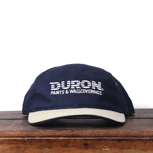 May include: A navy blue baseball cap with the white text "DURON PAINTS & WALLCOVERINGS" across the front. The brim and the band around the base of the cap are off-white.