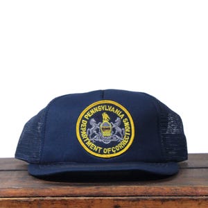 May include: A navy blue trucker hat with a mesh back and a curved brim. The front features a circular emblem with the text "PENNSYLVANIA DEPARTMENT OF CORRECTIONS" in gold lettering and a crest.