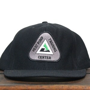 Puede incluir: Gorra de béisbol negra con un parche triangular gris que dice "Heat Pump Training Center" en texto blanco.