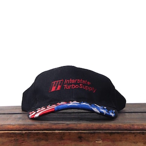 Peut inclure: Casquette de baseball noire avec le texte "Interstate Turbo Supply" brodé en rouge. La visière présente un motif d'étoiles rouges, blanches et bleues. Un accessoire de tête.