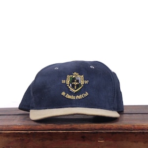 Puede incluir: Una gorra de béisbol azul marino con visera color canela. La parte frontal presenta un escudo bordado con una cruz, los números 18 y 97 a los lados y el texto St. Davids Golf Club en hilo dorado. La gorra tiene una corona estructurada.
