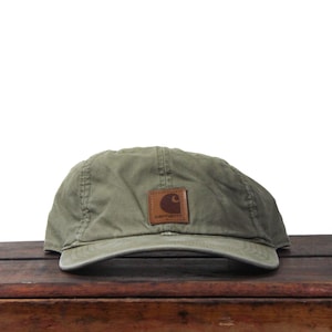 Puede incluir: Gorra de béisbol verde oliva con visera curva. Un parche de cuero marrón con el logotipo de Carhartt está cosido en la parte delantera. La gorra está hecha de una tela duradera y ligeramente desgastada.