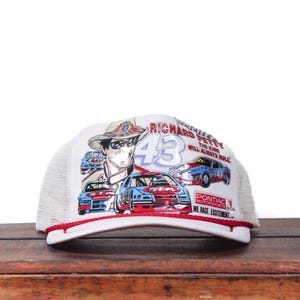May include: White trucker hat featuring a graphic of Richard Petty, the number 43, and race cars. The hat has a red brim and the text "RICHARD PETTY THE KING WILL ALWAYS RULE".