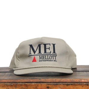 May include: A beige baseball cap with a black embroidered logo that reads "MEI MELLOTT ENTERPRISES INC." with a red triangle above the text.