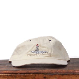 May include: A cream-coloured baseball cap with an embroidered lighthouse design on a rocky outcrop. The text "Peggy's Cove NS" is embroidered below. The cap has a curved peak and a fabric strap for adjustment.