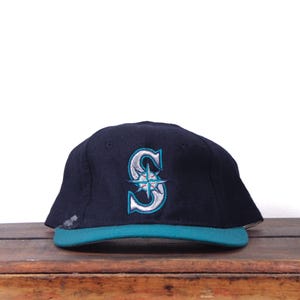 May include: A navy blue baseball cap with a teal brim. The cap features a white and teal embroidered "S" logo with a compass rose design. The cap is sitting on a wooden surface.