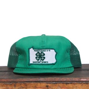 May include: Green and white Pennsylvania 4-H Shooting Sports trucker hat. The hat features a green front panel and bill, with a white patch displaying the 4-H clover and text. The sides and back are green mesh.