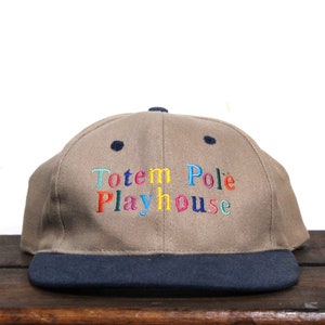 Peut inclure: Une casquette de baseball beige et bleu marine avec le texte "Totem Pole Playhouse" brodé en arc-en-ciel.