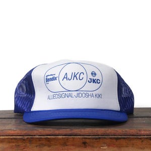 May include: A blue and white trucker hat featuring the text "AJKC" and "AlliedSignal-Jidosha Kiki". The hat has a blue mesh back and a blue brim.