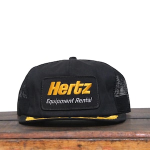 Könnte beinhalten: Schwarze Trucker-Cap mit Mesh-Rückseite und gelbem, gesticktem Rand. Auf der Vorderseite befindet sich ein rechteckiger Aufnäher mit dem Wort "Hertz" in Gelb und "Equipment Rental" darunter.