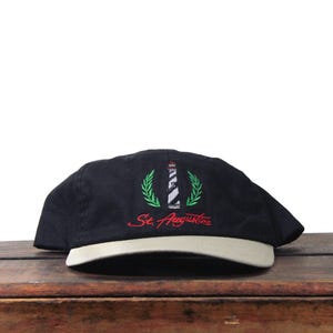 May include: Black baseball cap with a white brim. The cap features an embroidered design of a lighthouse, green laurel leaves, and the text "St. Augustine" in red.