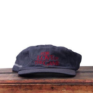 May include: Navy blue baseball cap with the embroidered text "FOR LOVE OF THE GAME" in red. The cap is sitting on a wooden surface. The cap also has text on the side.