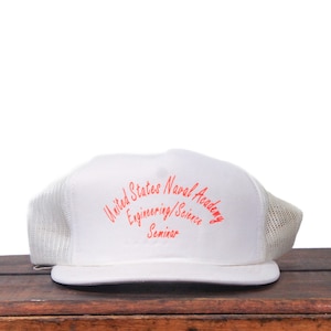 May include: White trucker cap with a mesh back and a curved peak. The front of the cap has the text "United States Naval Academy Engineering/Science Seminar" in red script. The cap is on a wooden surface.