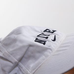 Puede incluir: Gorra de b&eacute;isbol blanca con la palabra "NIKE" bordada en negro en la parte delantera. El logotipo de Nike swoosh tambi&eacute;n est&aacute; bordado en negro. La gorra tiene una visera curva y un cierre de correa de tela.