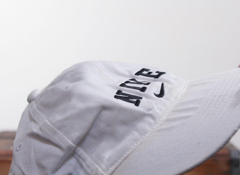 Puede incluir: Gorra de b&eacute;isbol blanca con la palabra "NIKE" bordada en negro en la parte delantera. El logotipo de Nike swoosh tambi&eacute;n est&aacute; bordado en negro. La gorra tiene una visera curva y un cierre de correa de tela.