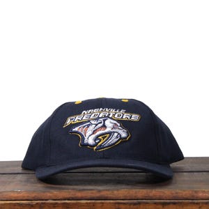 Może przedstawiać: Granatowa czapka bejsbolowa z haftowanym logo Nashville Predators. Logo przedstawia tygrysa szablozębnego w kolorze białym, żółtym i czarnym. Tekst "NASHVILLE PREDATORS" jest wygięty nad logo w kolorze białym.