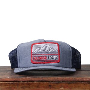 May include: A grey trucker cap with a black mesh back and a rectangular patch. The patch displays a mountain scene with the text "RAINIER LIGHT" in red.