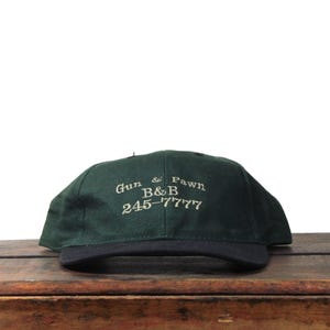 Może przedstawiać: Ciemnozielona czapka bejsbolowa z czarnym daszkiem. Na czapce znajduje się biały haft z napisem "Gun & Pawn B&B 245-7777". Czapka leży na drewnianej powierzchni.