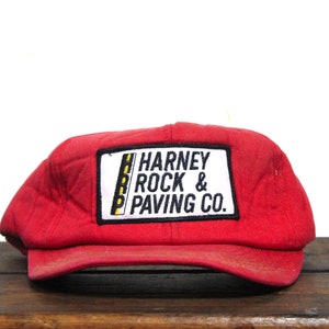 以下が含まれることがあります： 「Harney Rock & Paving Co.」と書かれた黒と白のパッチが付いた赤い野球帽