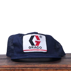May include: A navy blue trucker cap with a mesh back and a rectangular patch. The patch displays the word "GRACO" in white, with a black and red logo. The cap is on a wooden surface.