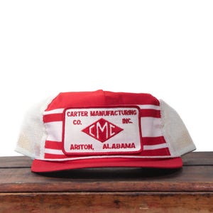 Könnte beinhalten: Rote und weiße Trucker-Mütze mit einem rechteckigen Aufnäher mit der Aufschrift "CARTER MANUFACTURING CO. CMC ARITON, ALABAMA". Die Mütze hat einen Mesh-Rücken und einen gebogenen Schirm.