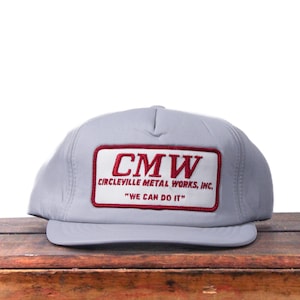 May include: A pale grey baseball cap with a flat peak. The cap has a rectangular patch with the letters "CMW" in red, and the words "CIRCLEVILLE METAL WORKS, INC." and "WE CAN DO IT" in red.