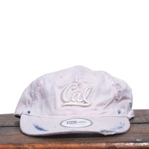 May include: A pink baseball cap with a white "Cal" logo embroidered on the front. The cap has a distressed look with some fraying around the edges.